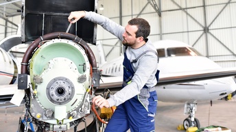 Person in blauer Arbeitslatzhose schraubt an Triebwerk eines Flugzeuges in Hangar, im Hintergrund verschwommen geparktes Flugzeug