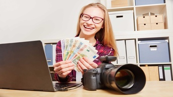 Eine Person sitzt im Büro vor Laptop mit Spiegelreflexkamera am Schreibtisch und EuroScheinen in der Hand.