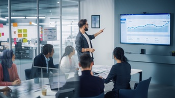 Mehrere Personen in Besprechungszimmer um Tisch sitzend, eine Person stehend deutet auf Monitor an Wand, auf dem statistische Kurven zu sehen sind