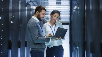 Zwei Personen stehen vor Servern und blicken auf aufgeklapptes Notebook, das eine Person in Händen hält