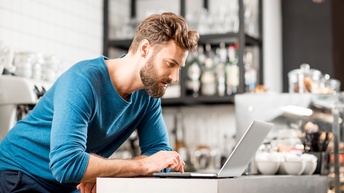 Person mit Bart in blauem Pullover über Laptop gebeugt, im Hintergrund Utensilien einer Bar