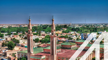 Stadtansicht von Nouakchott: Zentral rosafarbene Moschee mit grünen Dächern und zwei parallel gesetzten Minaretten, im Hintergrund niedrige Häuser und Bäume unter blauem Himmel.