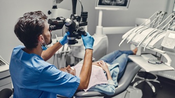 Person in blauer Arbeitskleidung mit Gummihandschuhen lugt durch Apparatur, die Licht auf darunter in Zahnarztstuhl liegender Person wirft, im Hintergrund zahnärztliche Geräte