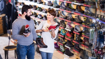 Zwei Personen mit Sport- und Wanderschuhen in Händen blicken einander an, im Hintergrund Regal mit Schuhen