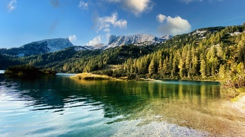 Im Vordergrund ist ein Gewässer. Im Hintergrund ein Gebirge mit Wald. Sie spiegeln sich im Wasser des Gewässers. Die Gipfel der Berge sind teilweise schneebedeckt.