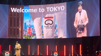 Eine Person steht auf einer Bühne. Hinter der Person ist eine große Leinwand, auf der rechts die Person ist und links daneben Welcome to Tokyo steht