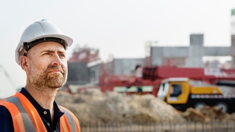 Person mit weißem Helm und oranger Schutzweste steht im Vordergrund einer Baustelle