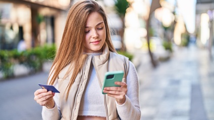 Eine junge Person in Pullover, Jeans und Gilets steht auf der Straße und blickt in ihr Smartphone, das sie in der linken Hand hält. In der rechten Hand hält sie eine Chipkarte