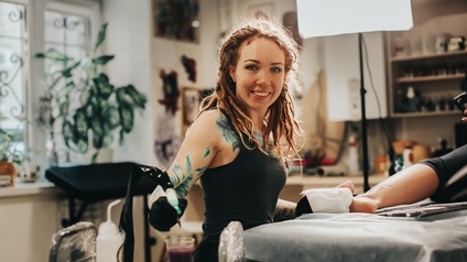 Lächelnde Person mit Tattoos auf Armen und Oberkörper sitzt Tisch, auf der andere Person im Ausschnitt liegt und taucht Tätowiermaschine in ein Behältnis mit Farbe