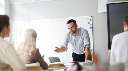 Person steht in einem Zimmer mit Whiteboard, großem Bildschirm und zeigt auf eine weitere Person, die auf einem Tisch mit Büchern sitzt währendweitere Personen aufmerksam zuhören