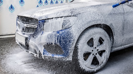 Blaues Auto steht in einer Waschanlage ist von weißem Schaum bedeckt und wird von einem Wasserstrahl gesäubert