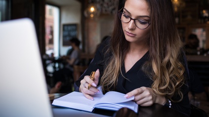 Brillen tragende Person mit langen dunklen Haaren blickt in vor ihr liegendes Kalenderbuch und hält Kugelschreiber in einer Hand, im Hintergrund verschwommen Lokal mit weiteren Personen