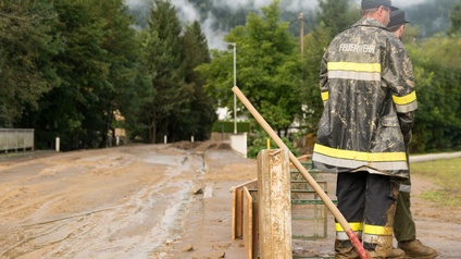 Zwei Personen in Rückenansicht in Schutzkleidung mit der Aufschrift Feuerwehr stehen neben wasserverschlammter Straße, die von Bäumen umsäumt ist, neben den Personen lehnt eine Schaufel an Holzbalken.