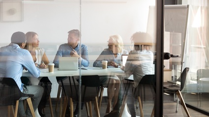 Personen in Businesskleidung sitzen an einem Besprechungstisch mit Laptop und weiteren Unterlagen in einem gläsernen Büro