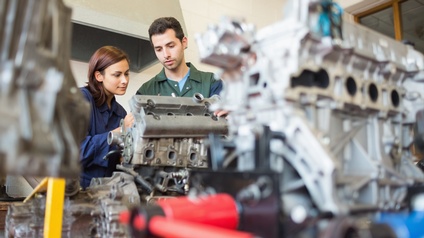 Zwei Personen inspizieren Automotor, im Vordergrund verschwommen weitere Motorenteile