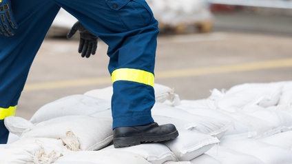 Nahaufnahme des rechten Beins einer Person in einem schwarzen Schuh mit dicker Sohle. Der Fuß steht auf weißen übereinander gestapelten Säcken. Im Hintergrund verschwommen ist eine asphaltierte Straße.