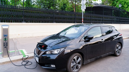 Schwarzes Elektroauto in Seitenansicht an Stromzapfsäule mit Kabel verbunden Strom tankend, im Hintergrund weiße Mauer mit schwarzem Zaun, durch den Grün von Pflanzen schimmert