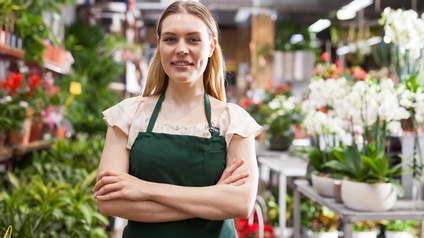 Portrait einer Person in grüner Schürze, die Arme verschränkt vor Körper hält, im Hintergrund verschwommen Regale und Tische mit Blumen