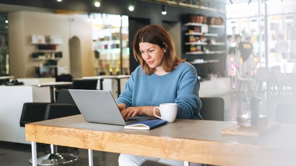 Person sitzt an hohem Tisch in Kaffeehaus und blickt auf aufgeklappten Laptop