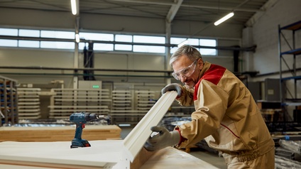 Person mit grauen kurzen Haaren und Bart sowie Arbeitskleidung und Brille steht in einer Werkstätte und hält eine Vorhangkarniese aus weißem Holz, daneben stehet ein Akkuschrauber