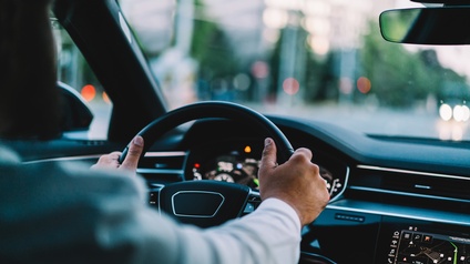 Blick von der Rückbank eines Autos auf zwei Hände einer Person auf einem Lenkrad in einem Auto auf einer Straße