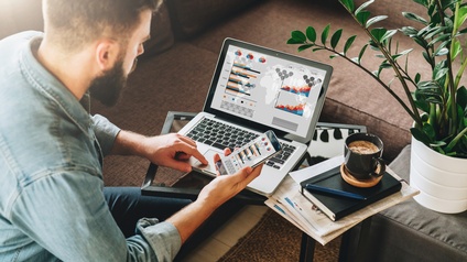 Person mit Hemd sitzt vor einem Laptop an einem Couchtisch und hält sein Smartphone in der Hand während er Statistiken auf beiden Geräten in einer Wohnzimmersituation betrachtet, daneben liegen Zeitungen, ein Notizbuch sowie eine Tasse Kaffee und eine Grünpflanze