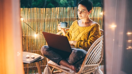 Person mit kurzen dunklen Haaren und gelbem Pullover sitzt mit einer Tasse und einem Laptop auf einem Balkon am Abend, dahinter erhellt eine Lichterkette die Atmosphäre