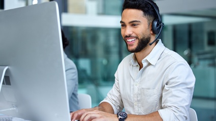 Eine lächelnde Person mit einem Headset sitzt in einem hellen Großraumbüro vor einem weißen Screen und schreibt etwas auf der Tastatur.