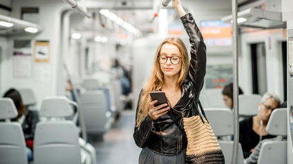 Eine Person steht in einem Zugabteil und hält sich mit der linken Hand an einer horizontalen Stange fest. In der rechten Hand hat sie ein Smartphone, auf das sie blickt. Im Hintergrund sitzen auf Leute vereinzelt auf Sitzen