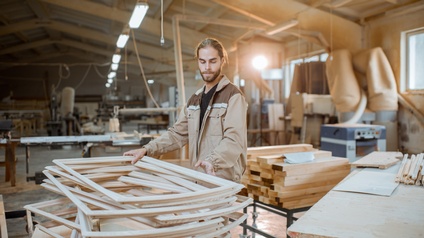 Person mit braunen geschlossenen Haaren und Bart sowie Arbeitsjacke steht bei Holzrahmen in einer Tischlerwerkstätte