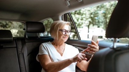 Pensionierte Person mit kurzen blonden Haaren, Brille und weißer Bluse sitzt auf der Rückbank eines Personenwagens und bedient ein Smartphone