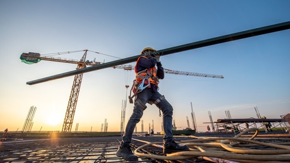 Person ich Schutzkleidung trägt großen Metallbalken über Baugrund, im Hintergrund Kran im Sonnenuntergang