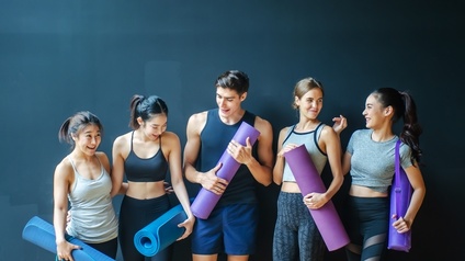 Mehrere lächelnde Personen in Sportkleidung stehen nebeneinander vor dunkelblauer Wand und halten violette und blaue zusammengerollte Gymnastikmatten