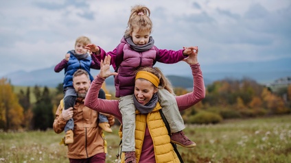 Zwei Erwachsene und zwei Kinder in Wanderkleidung gehen über eine Wiese. Je eine erwachsene Person trägt ein Kind auf ihren Schultern