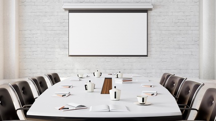 Weißer Besprechungstisch mit acht schwarzen Stühlen, auf dem Kaffeetassen, Kannen und Arbeitsutensilien liegen, im Hintergrund auf weißer Ziegelwand Leinwand hängend