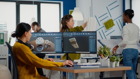 Person mit Kopfhörern um den Hals sitz an Schreibtisch vor zwei großen Monitoren auf denen Renderings von Zahnrädern und Bauteilen zu sehen sind, im Hintergrund weitere Personen stehend in Büroraum