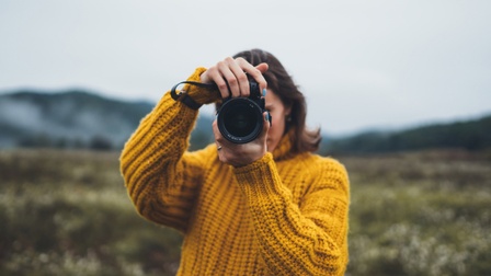 junge Frau in gelbem Strickpullover steht in nördlicher Landschaft und hält eine Kamera vor dem Gesicht.