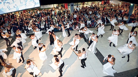 Blick auf eine Maturaball-Polonaise mit vielen Tanzpaaren - Männer im schwarzen Anzug, Frauen im weißen Kleid 