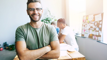 Person mit kurzen dunklen Haaren und Brille sowie grünem Shirt steht mit verschränkten Armen vor einem Besprechungstisch an dem weitere Personen sitzen, im Hintergrund steht eine Pinnwand mit vielen kleinen angehefteten Zetteln