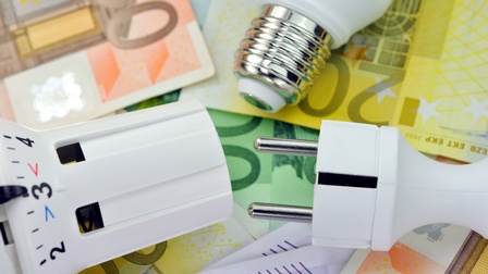 Nahaufnahme mehrerer Euro-Geldscheine in den Farben orange, grün, lila und gelb. Auf den Geldscheinen liegen eine Glühbirne, ein Thermostat und ein Stecker.