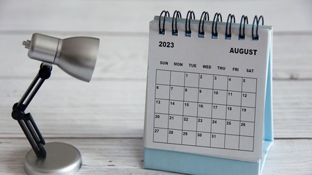 Stehkalender mit Kalenderblatt von August 2023 steht gemeinsam mit einer Miniatur-Schreibtischlampe auf einem weißen Holzuntergrund