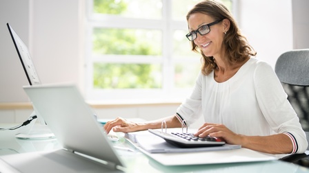 Person mit Brille sitzt an einem Schreibtisch und arbeitet mit einem Laptop