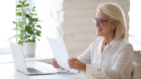 An Schreibtisch sitzende lächelnde Person mit Brillen blickt auf Blatt in Händen, am Tisch aufgeklappter Laptop, im Hintergrund Zimmerpflanze