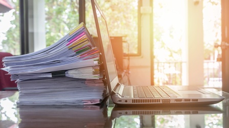 Dokumentenstapel an dem aufgeklapptes Notebook lehnt, beides spiegelt sich in Oberfläche, im Hintergrund verschwommen Fenster., durch die Sonnenlicht einfällt