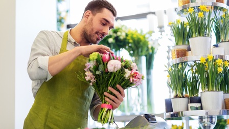 Bärtige Person in grüner Schürze hält Blumenstrauß in Händen, blickt darauf und greift danach, ringsum weitere Pflanzen stehend