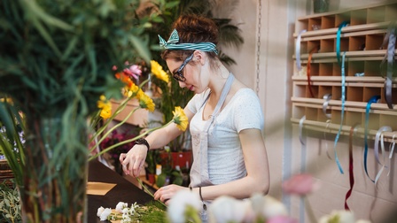 Person in Schürze mit gestreiftem Haarband und Brillen legt auf Tisch Blumen aufeinander, ringsum weitere Pflanzen, auf der Wand hinter Person Regal mit Geschenksbändern