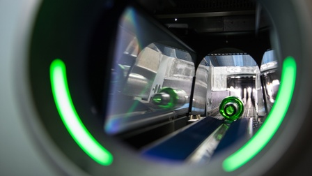 Detailansicht einer Rollbandes eines Getränkerückgabeautomaten, im Vordergrund verschwommen grüne Lichter, auf Rollband grüne Glasflasche liegend