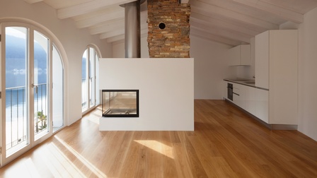 In einem hellen Raum sind links zwei viertürige Fenster Richtung eines Sees. In der Mitte ist ein Ofen mit Kamin. Rechts befindet sich eine weiße Küche.