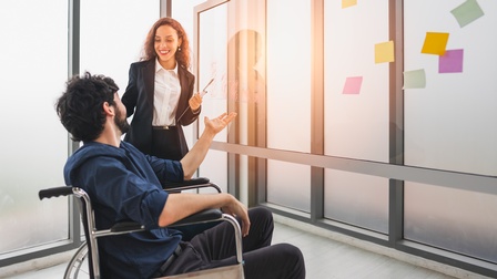 Person in Rollstuhl unterhält sich mit einer weiteren Person vor einer Glasfront mit Post-Its und Notizen in Businesskleidung