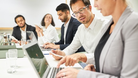 Mehrere Personen um Tisch sitzend: Fokus auf einer Person, die spricht und auf Tastatur neben ihr vor aufgeklapptem Laptop und Hände auf Tastatur sitzender weiterer Person tippt